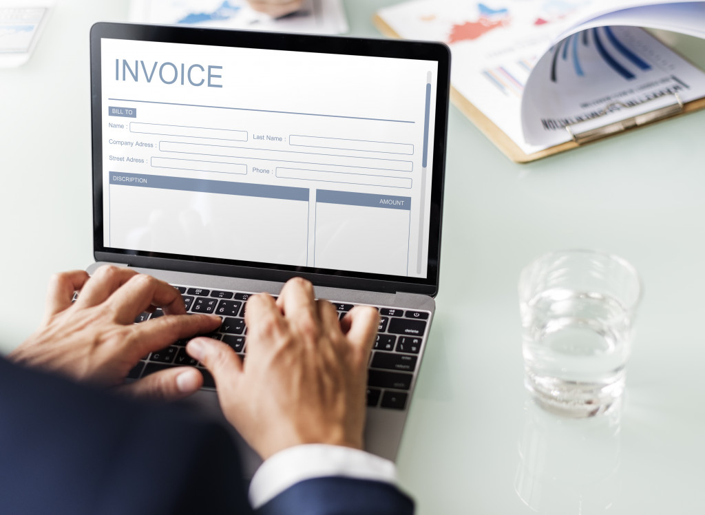 Person preparing an invoice using a laptop on a table with a glass of water and documents on it.